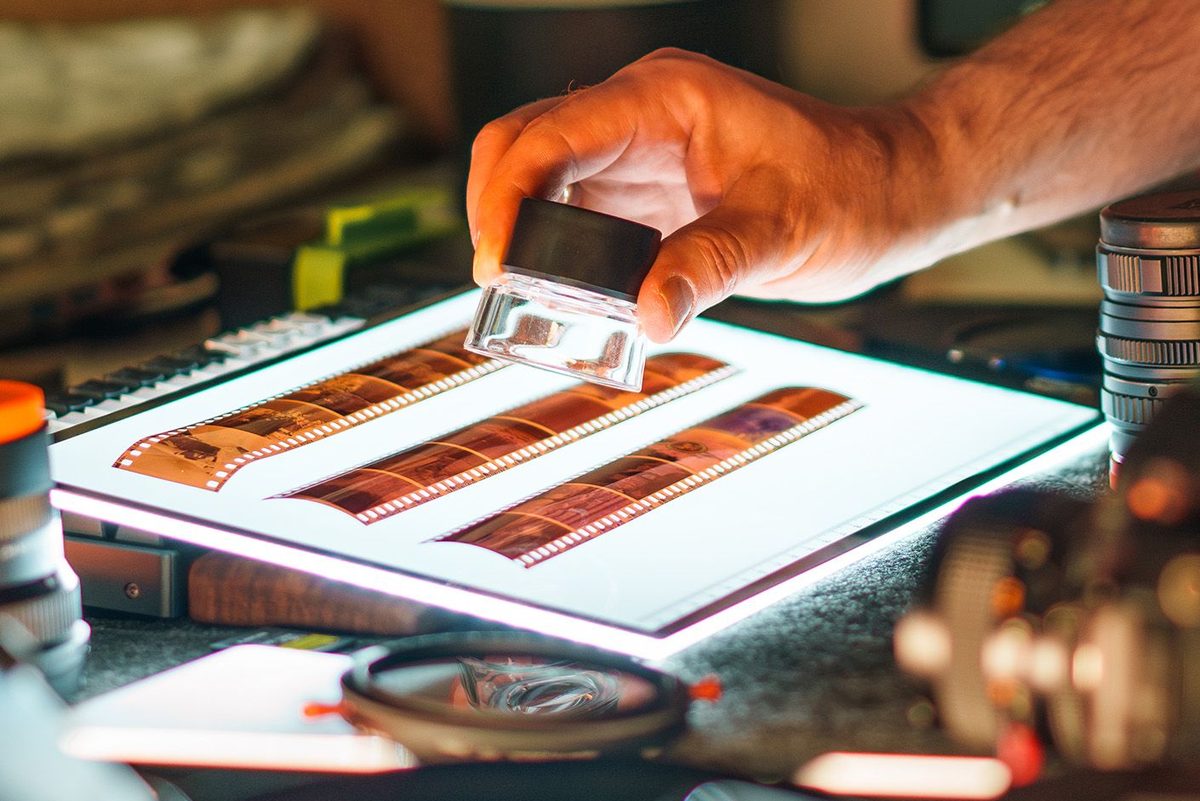 A hand holding a loupe over strips of film negatives on a lit light table
