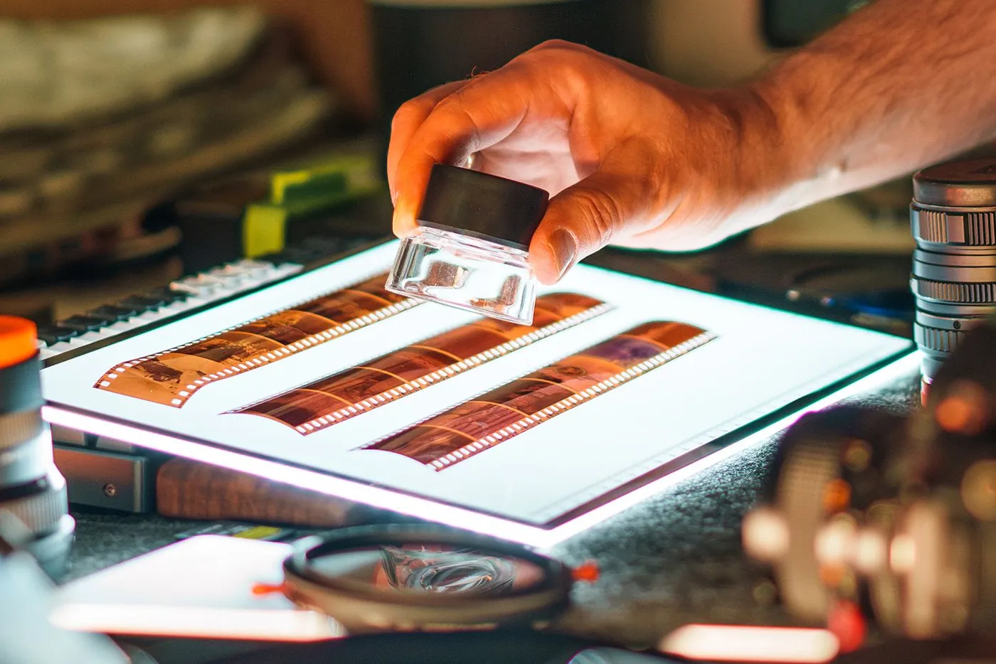 A hand holding a loupe over strips of film negatives on a lit light table
