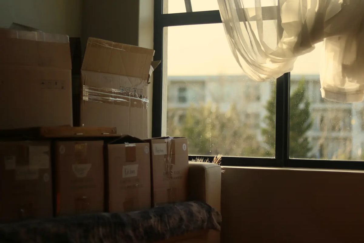 Taped cardboard moving boxes stacked in a corner beside a sunlit window with a sheer curtain