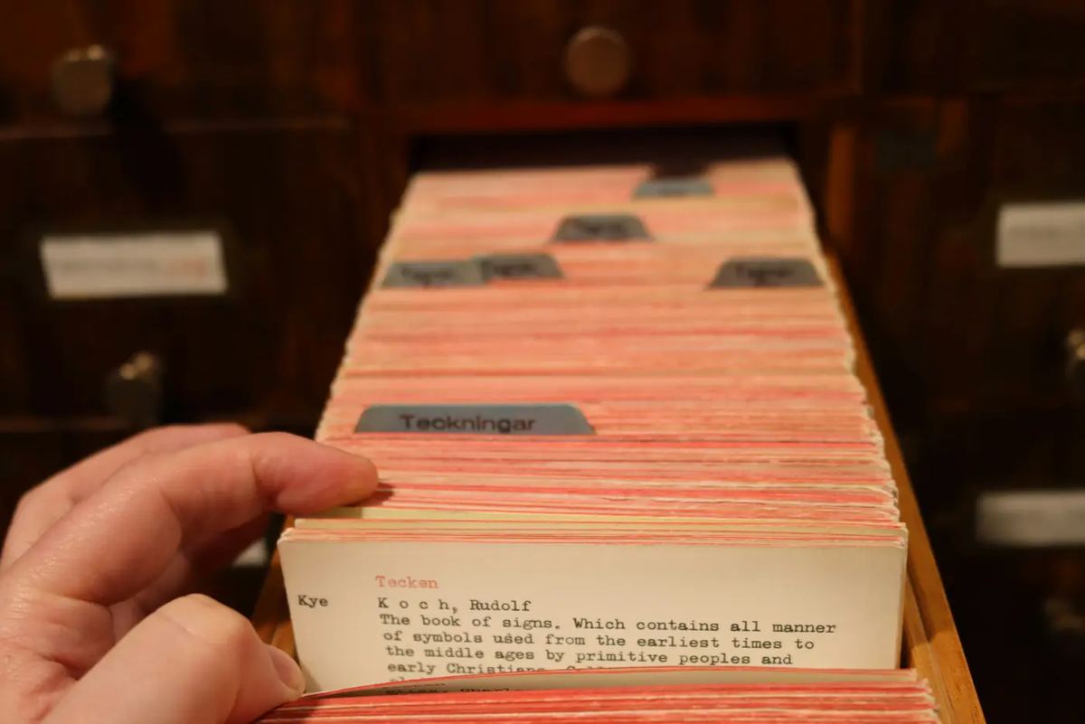 A hand pulling a single card out of an open wooden library card catalogue drawer