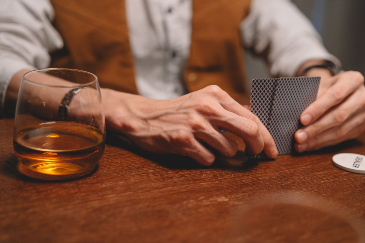 Two hands holding playing cards over a dark wooden table, with a glass of whisky beside them