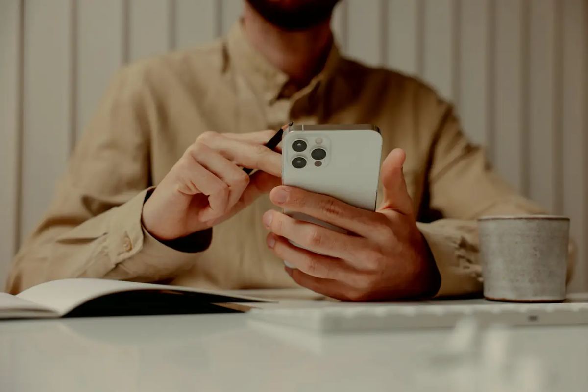 Someone at a pale desk tapping a phone held in one hand, an open notebook and a ceramic mug beside them